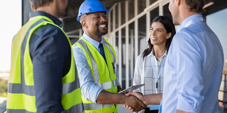Man in dress shirt shakes hands with a contractor in a hard hat and high viz vest while two other people look on