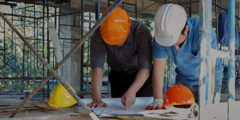 Two contractors reviewing blueprints under a scaffolding.