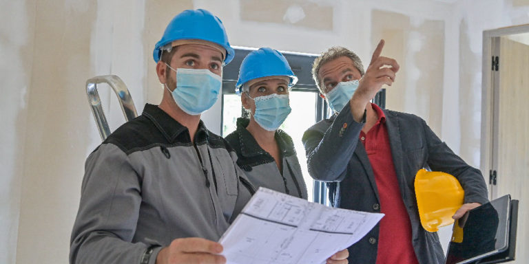 Three people wearing masks and hard hats look up at something off screen.