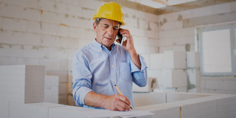 Man in collared shirt and hard hat talking on a phone while standing inside of a building under construction.