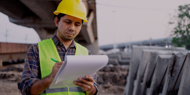 Man in yellow hard hat and green vest reviews a document on a jobsite.