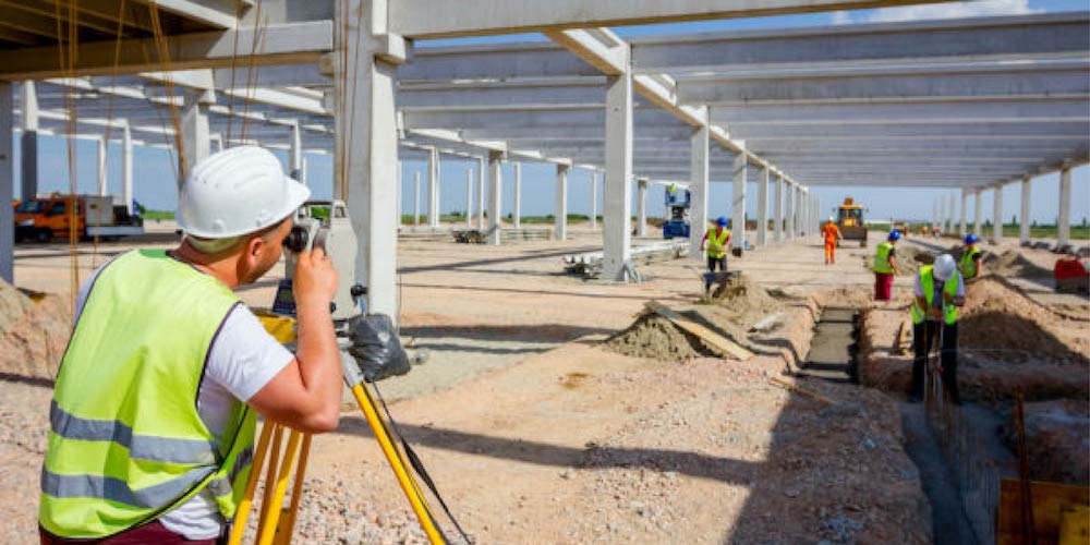 Man in hard hat and vest surveys a construction site with workers and equipment.