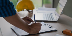 Close up of hand holding pen reviewing blueprints with computer and hard hat in the background.