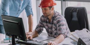 Man in hard hat looking at a computer screen while another man looks on.