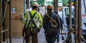 Two construction workers wearing safety harnesses walk under scaffolding in a city.