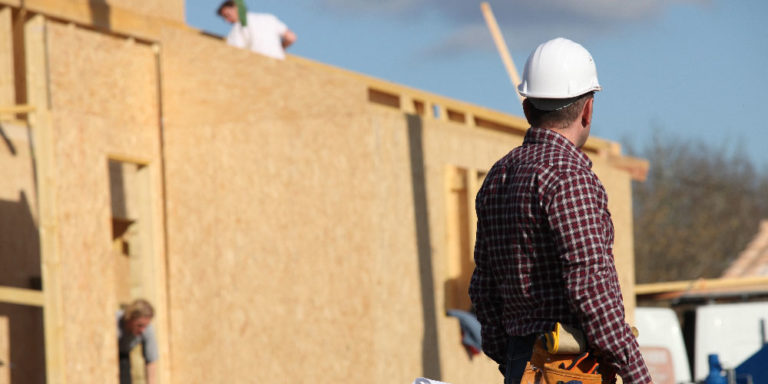 Construction worker with back to camera walks in front of a building being framed plywood.