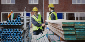 Two construction workers shake hands behind stacks of building materials.