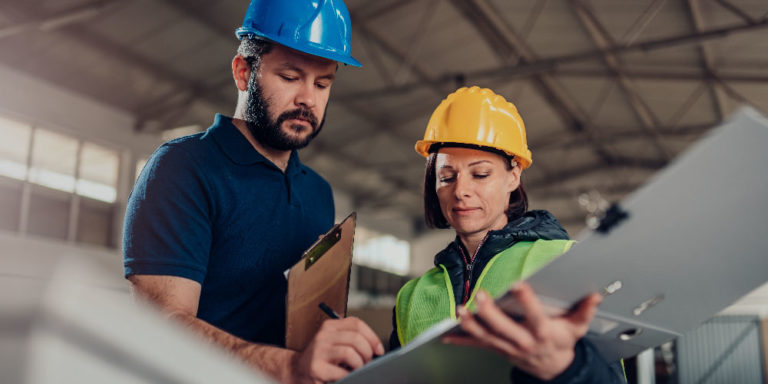 Man and woman wearing hard hats and reviewing a document inside of a warehouse.
