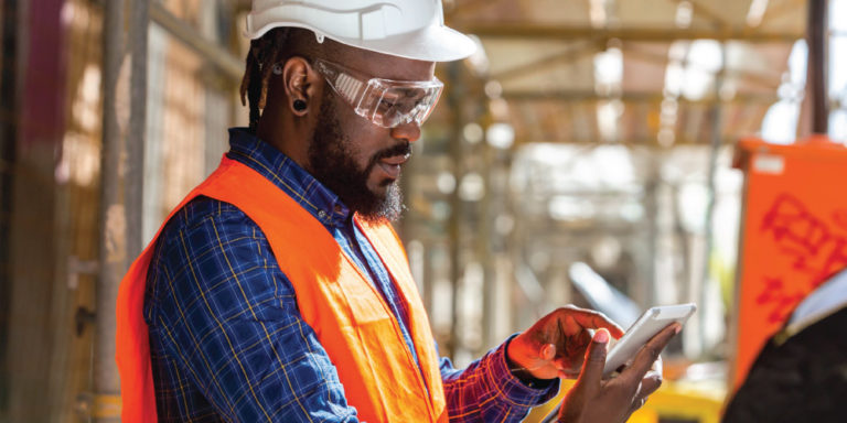 Man in hard hat and high visibility vest looks at tablet.