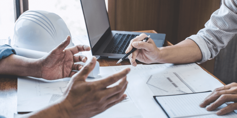 Photo of 2 people's hands as they discuss change order documents on a desk with a laptop and hard hat pictured.