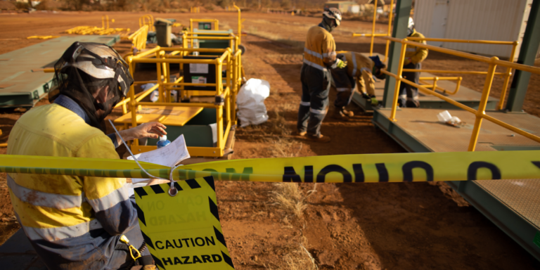 Construction accidents: photo of caution tape on construction site with 2 workers wearing protective equipment working inside and checking documents.