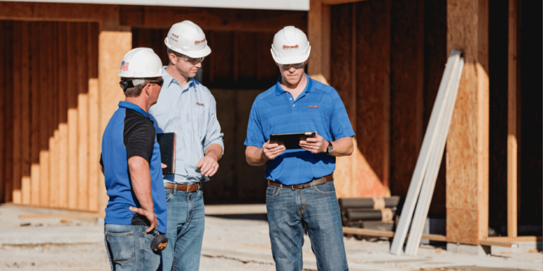 3 workers stand in the middle of a construction site wearing hardhats; the one on the far right is reviewing a tablet.