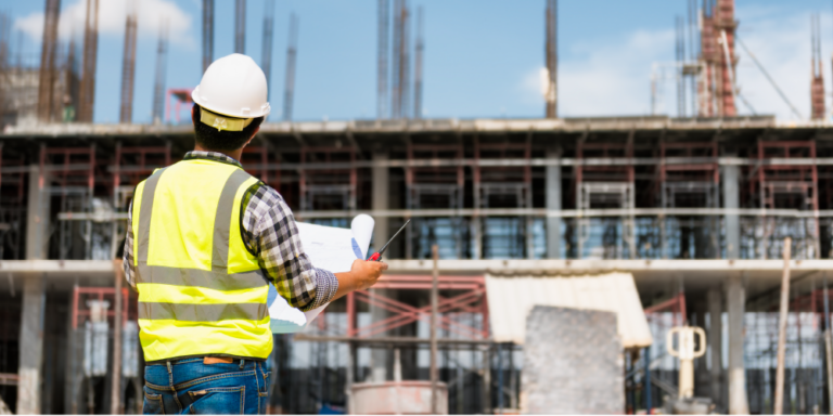 Contractor in high viz vest and hard hat holds drawings and looks at a building under construction