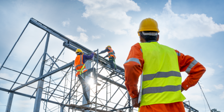 Three workers on a jobsite all wearing PPE including reflective vests and hardhats and harnesses. One worker is supervising two others who are climbing scaffolding.