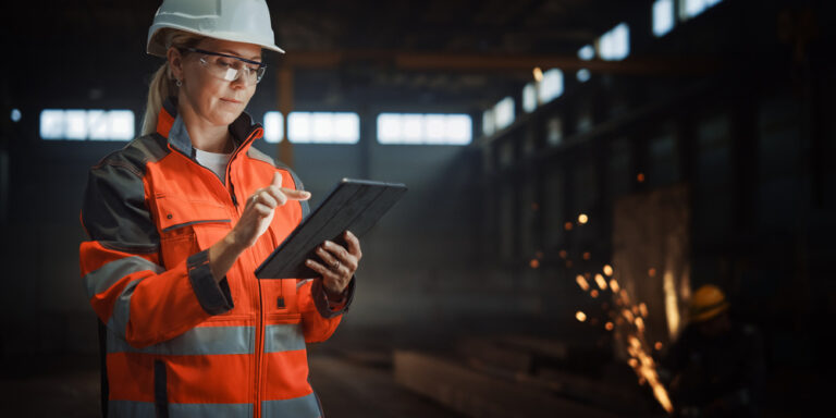 Female engineer in hard hat and high viz vest reviews BIM drawings on a tablet.
