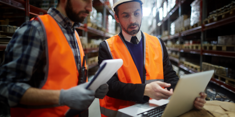 2 contractors in a materials warehouse looking at construction procurement lists on a clipboard and laptop