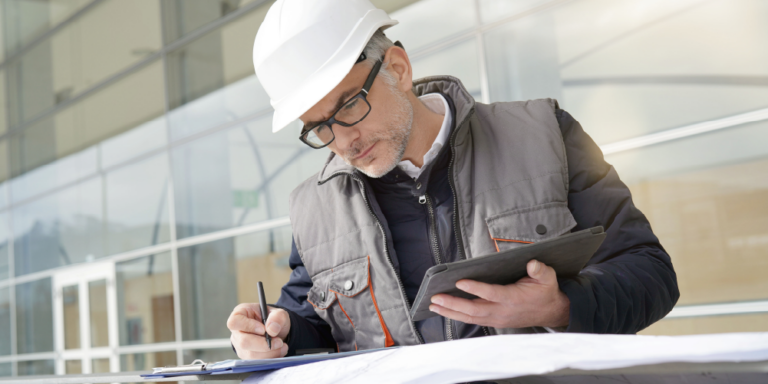 Photo of a construction project manager in a hard hat looking at a tablet and documents