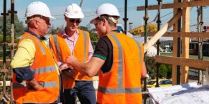 Three construction professionals wearing safety vests and hard hats on a jobsite discussing project details.