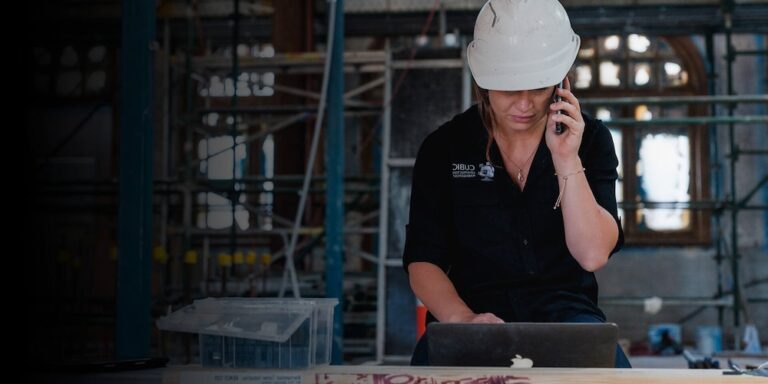 Woman wearing hard hat on a jobsite reviews documents on a laptop.