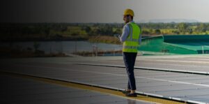 A man in a safety vest and hard hat holding a tablet walks along a field of solar panels.