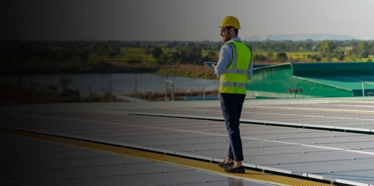 A man in a safety vest and hard hat holding a tablet walks along a field of solar panels.