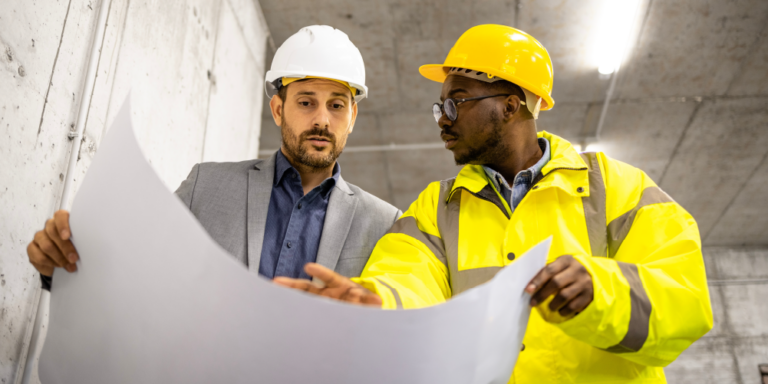 A man in formal dress and another man wearing safety gear review drawings on a construction jobsite.