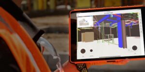 A person on a construction jobsite with a safety vest holds up a tablet displaying a 3D BIM model