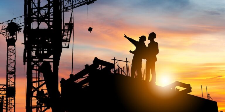 Two construction professionals surveying a site and pointing upward toward a crane, silhouetted by the setting sun.
