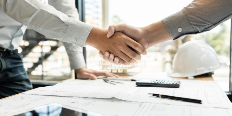 Closeup of handshake with construction documents and hard hat on a desk.