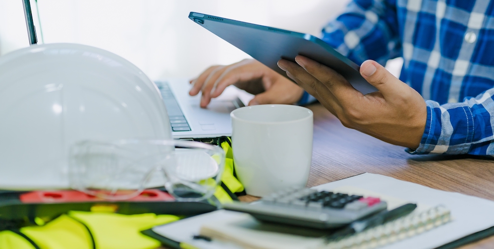 A construction estimator looking at a tablet with a hard hat and vest on the desk in front of them