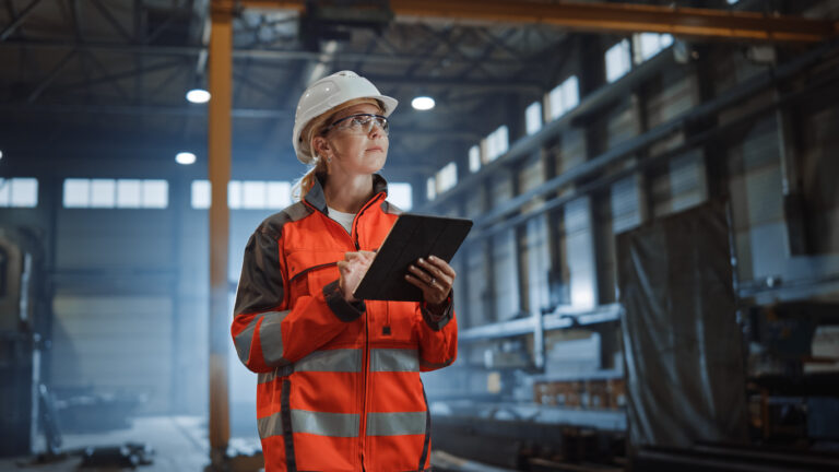 Professional heavy industry engineer worker wearing safety uniform and hard hat, using tablet computer in a metal manufacture warehouse.