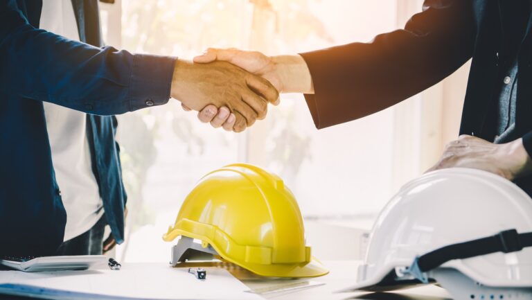 Construction partnering 2 construction professionals shaking hands over a hard hat and paperwork