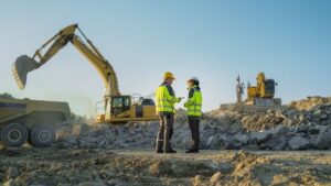 Two construction workers talking on a jobsite with an excavator in the background