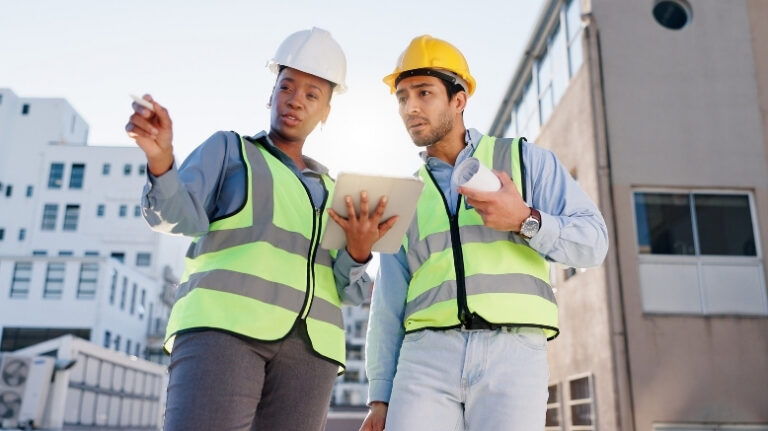 Two workers on a jobsite gesture toward something on-site that is reflected on a digital tablet