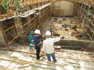An elevated view of a large building under renovation or construction. In the foreground, three people wearing hard hats — two in white helmets and one in a green helmet — stand on an elevated platform, appearing to discuss the project while gesturing toward the work area below. The site features extensive yellow scaffolding along the walls, scattered bricks and construction materials on the ground floor, and several workers in green high-visibility uniforms visible throughout. The structure appears to be a large interior space, possibly a historic building undergoing restoration, with arched architectural elements visible in the background.