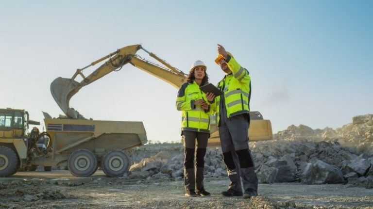 Two workers on a jobsite talking with an excavator behind them